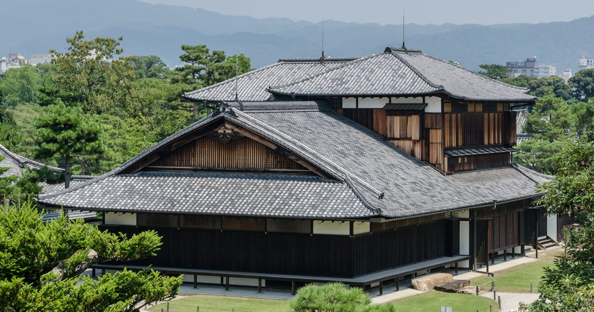 Der Honmaru-Palast des Nijo Castle, ein zweistöckiger Holzpavillon zwischen Kiefern, gesehen vom Sockel des Bergfrieds mit Kyotos Hügeln im Hintergrund